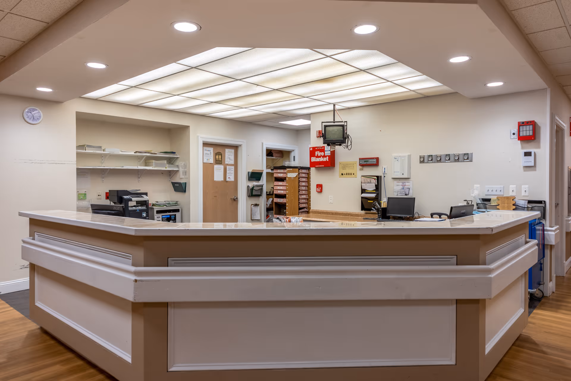 Reception desk area in a senior living facility with a large counter, computer monitors, office equipment, shelves with files, and a fire blanket mounted on the wall. The ceiling has bright fluorescent lighting panels.
