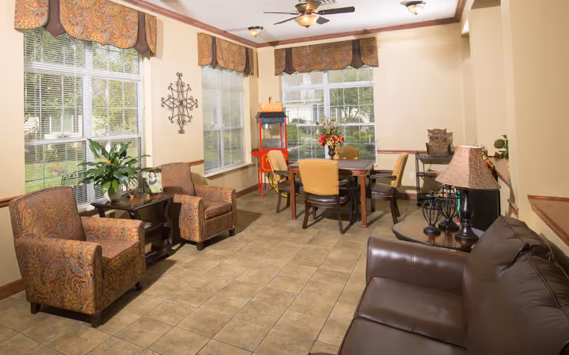 A cozy living room area with a brown leather sofa on the right, two patterned armchairs on the left, and a wooden table with four chairs in the background. The room has large windows with decorative valances, a ceiling fan with lights, and a popcorn machine near the windows. There are plants and decorative items on side tables, and the floor is tiled.