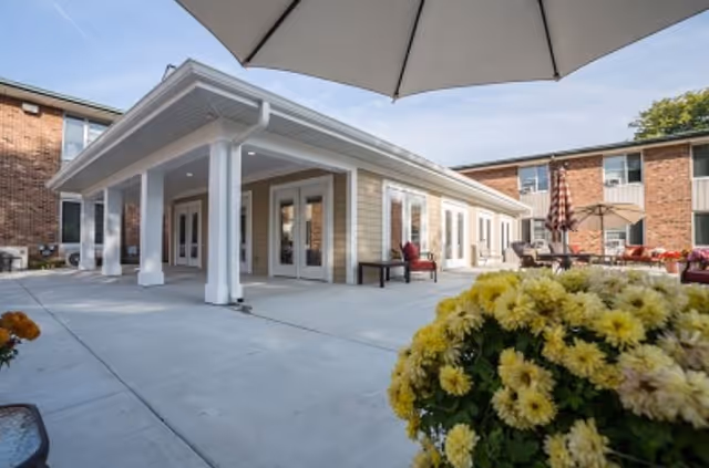 Courtyard patio with umbrellas, seating, and potted yellow flowers outside a low brick-and-siding senior living building.