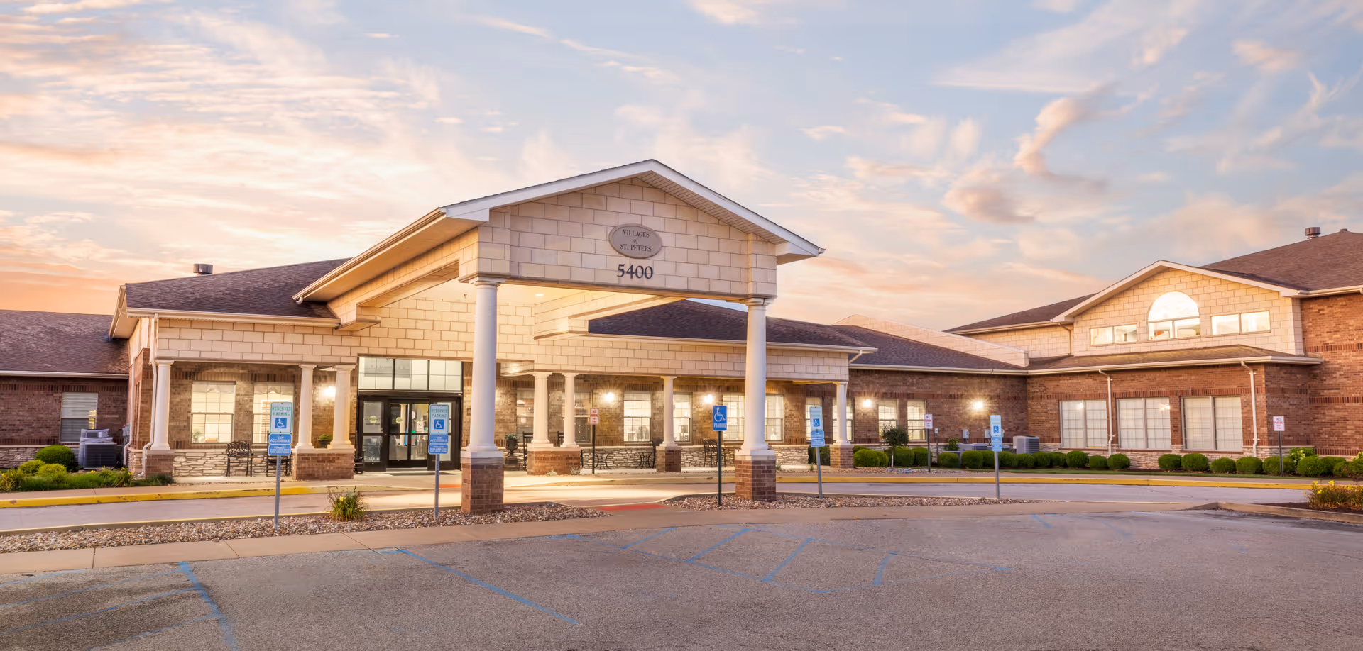 Front exterior view of the Villages of St. Peters facility building at sunset, featuring a covered entrance with columns, multiple windows, and a parking area with handicap parking spaces in front.