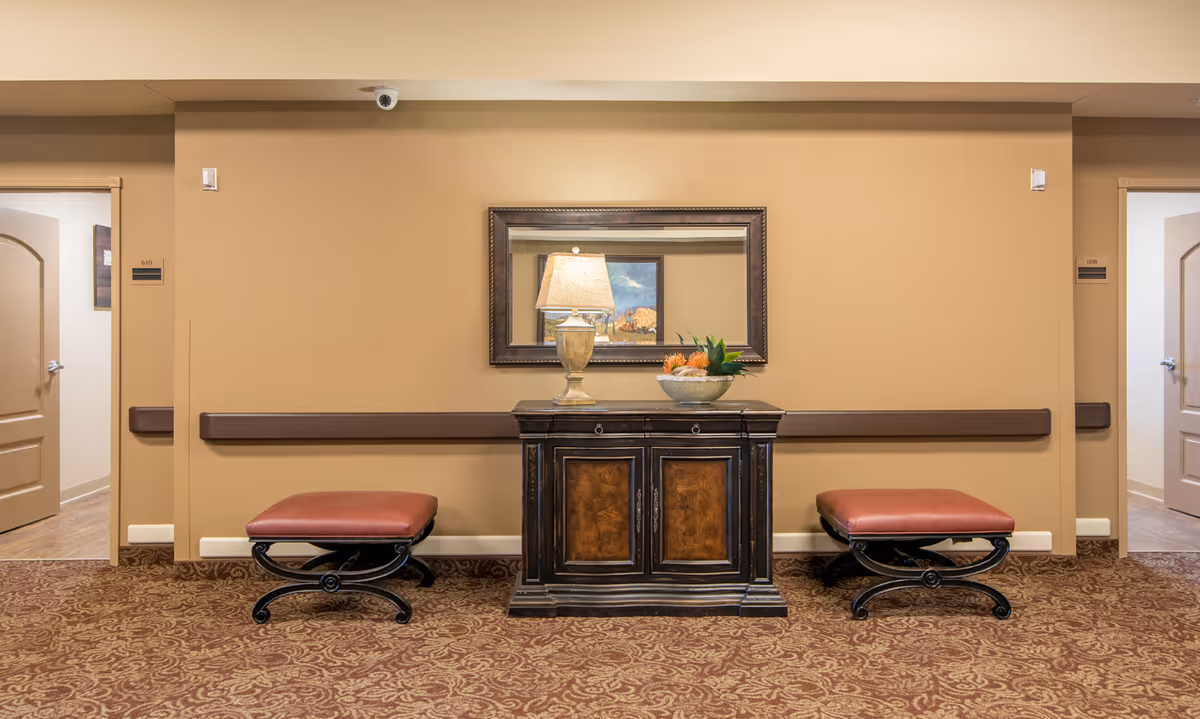 A hallway area in a senior living facility with beige walls and patterned carpet. There is a dark wooden cabinet with a lamp and a decorative bowl on top, flanked by two cushioned benches with red upholstery. A large framed mirror hangs above the cabinet, reflecting part of a painting. Two open doors lead to other rooms on either side of the hallway.