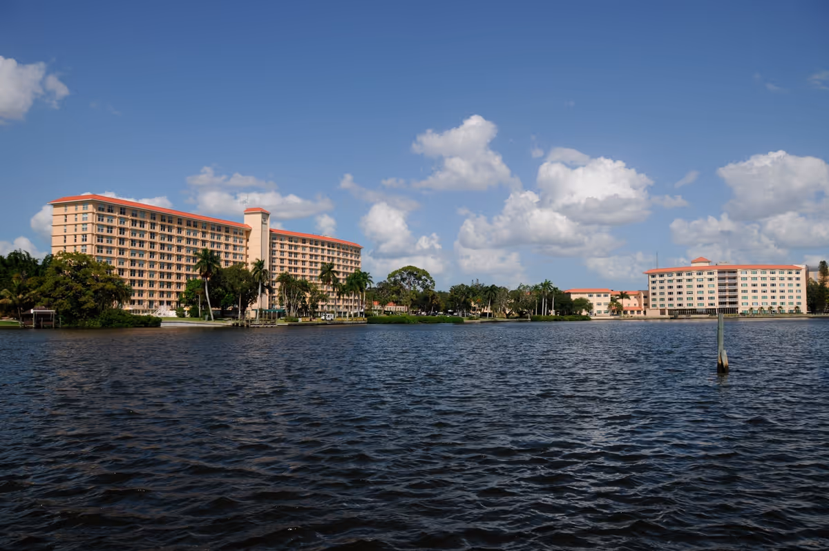 View of two large multi-story buildings with peach-colored walls and red roofs situated along a waterfront under a blue sky with scattered white clouds. Palm trees and other greenery are visible near the buildings.