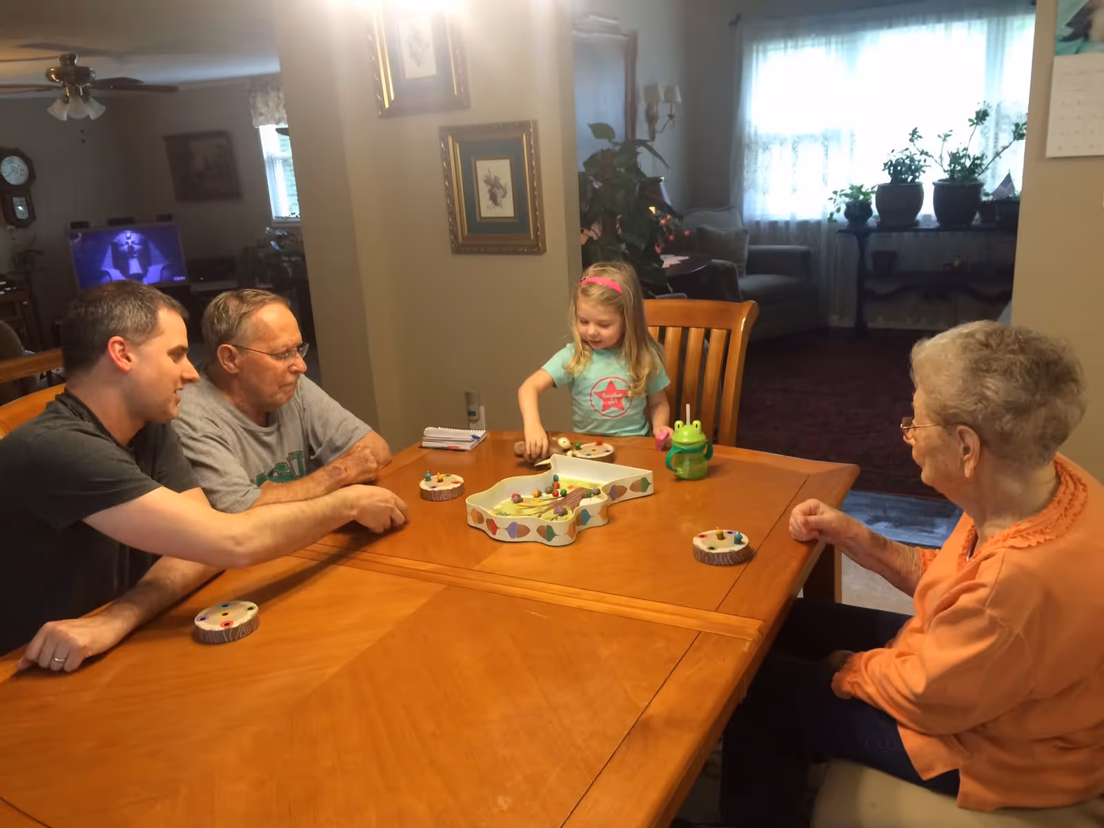 A young girl and three adults, two men and one elderly woman, sitting around a wooden dining table playing a board game in a cozy home setting with framed pictures on the wall and a window with sheer curtains in the background.