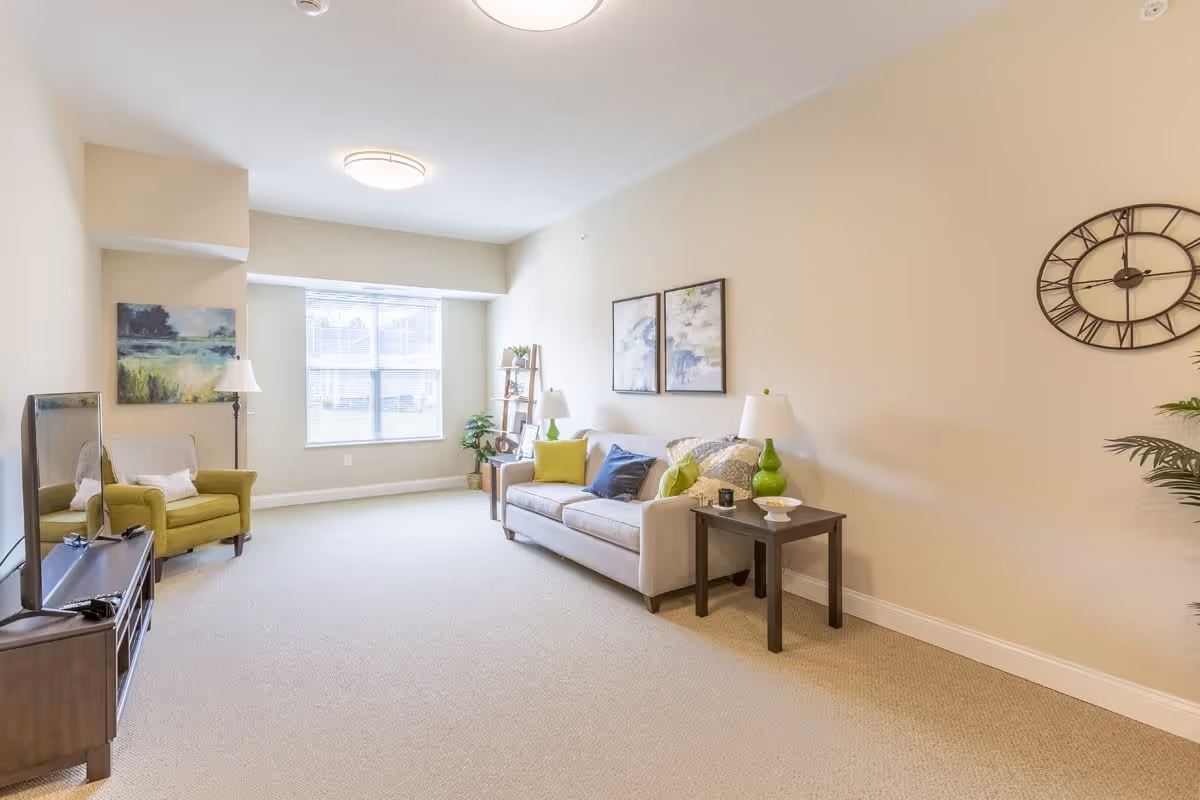 A bright living room with beige walls and carpet. The room features a beige sofa with colorful pillows, two side tables with lamps, a green armchair, a TV on a wooden stand, a large window letting in natural light, wall art, a decorative clock, and some plants.