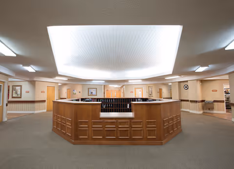 Reception lobby with a large wooden central desk under a recessed illuminated ceiling and surrounding corridors and doors.