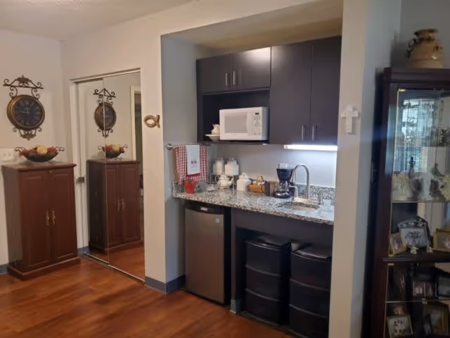 A small kitchenette area with dark upper cabinets, a granite countertop, a microwave, a coffee maker, and a small refrigerator. To the left, there is a wooden cabinet with a decorative clock and a bowl of fruit on top. A mirrored closet door is also visible. To the right, there is a glass display cabinet with various decorative items inside.
