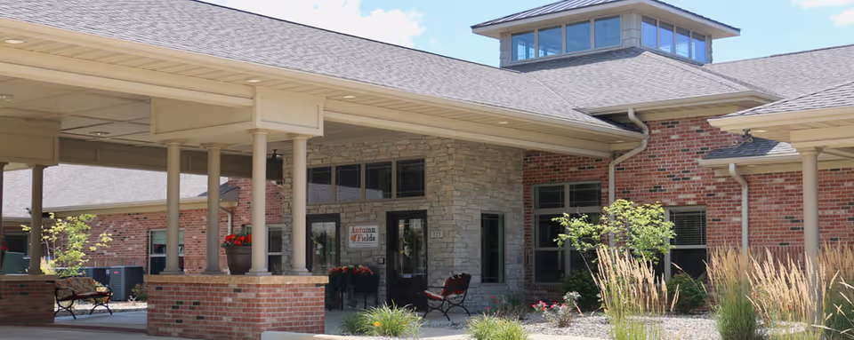 Exterior view of Autumn Fields Adult Community-Hoopeston showing a covered entrance with brick and stone walls, benches, plants, and a clear sky above.