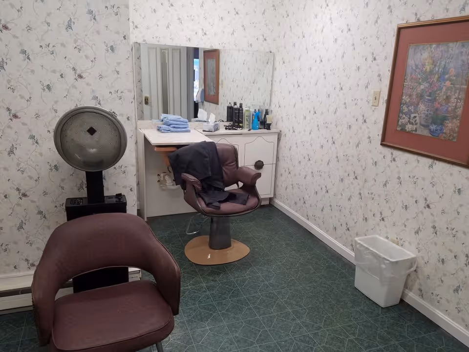 A small room with floral wallpaper and green tiled floor, featuring two brown salon chairs, a hair dryer, a vanity with a large mirror, various hair care products, folded towels, and a framed floral painting on the wall. There is also a white trash bin in the corner.