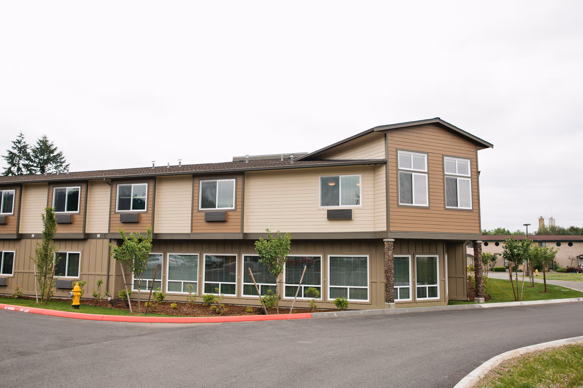 Exterior view of a two-story senior living facility building with beige and brown siding, multiple windows, small trees, and landscaping along the front. The sky is overcast and there is a paved driveway in front of the building.