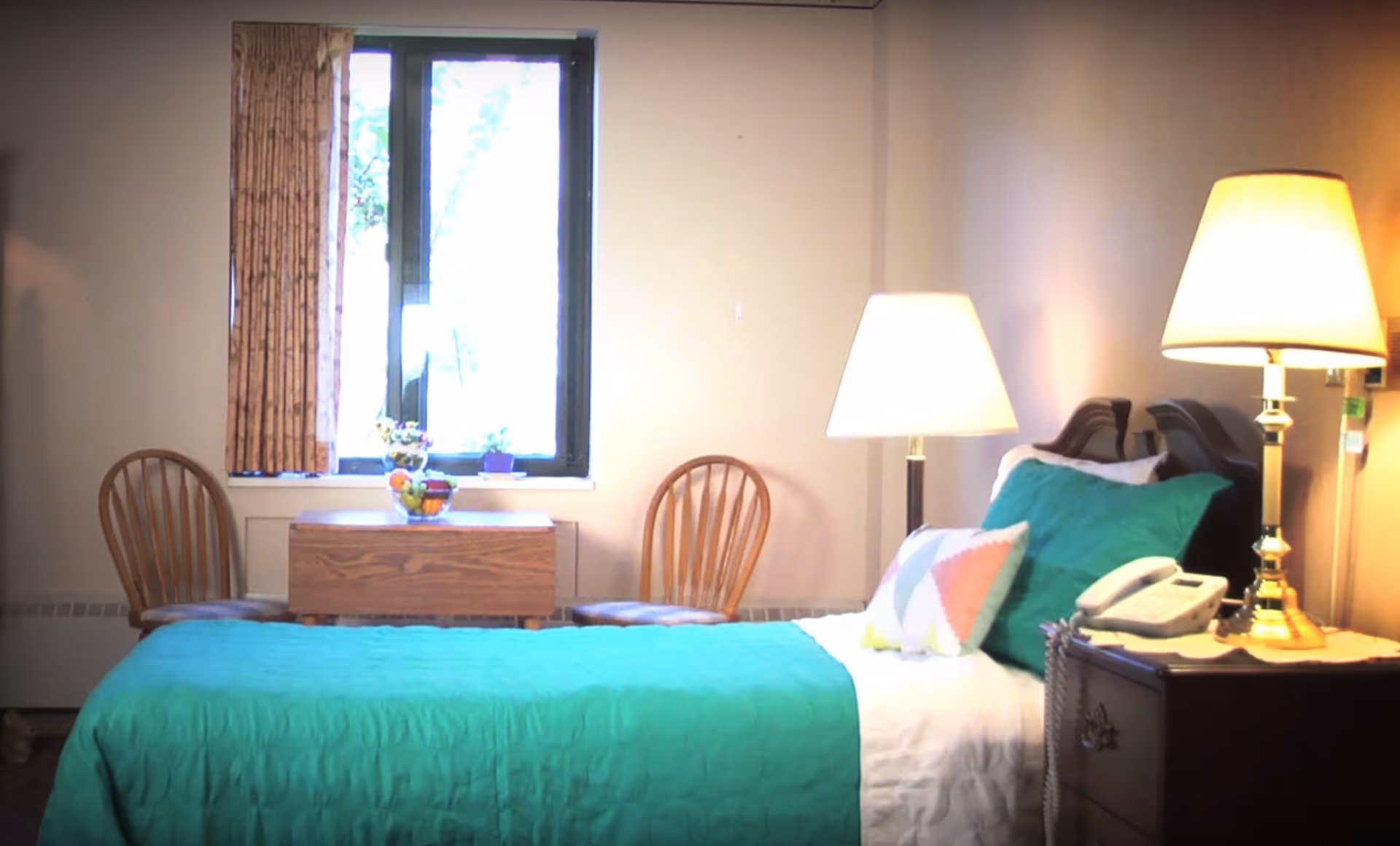 A bedroom with a single bed covered in a green quilt and a colorful pillow. There are two wooden chairs and a small wooden table with a bowl of fruit and a small plant in front of a window with patterned curtains. A nightstand beside the bed holds a telephone and a brass lamp with a white lampshade.