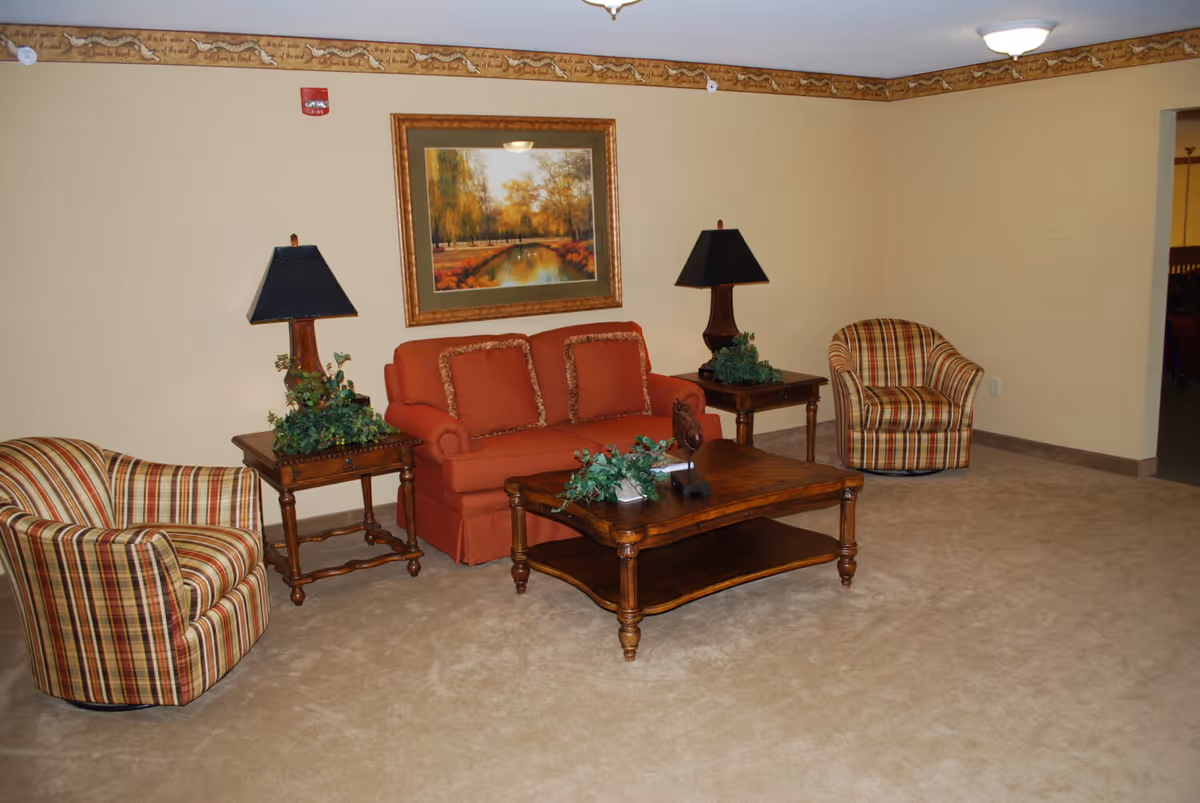 A living room area with a red sofa flanked by two wooden side tables, each with a black lampshade and green plant decoration. Two striped armchairs are positioned on either side of the sofa. A wooden coffee table with decorative greenery is in front of the sofa. The walls are beige with a decorative border near the ceiling, and a framed painting of a serene autumn landscape hangs above the sofa.