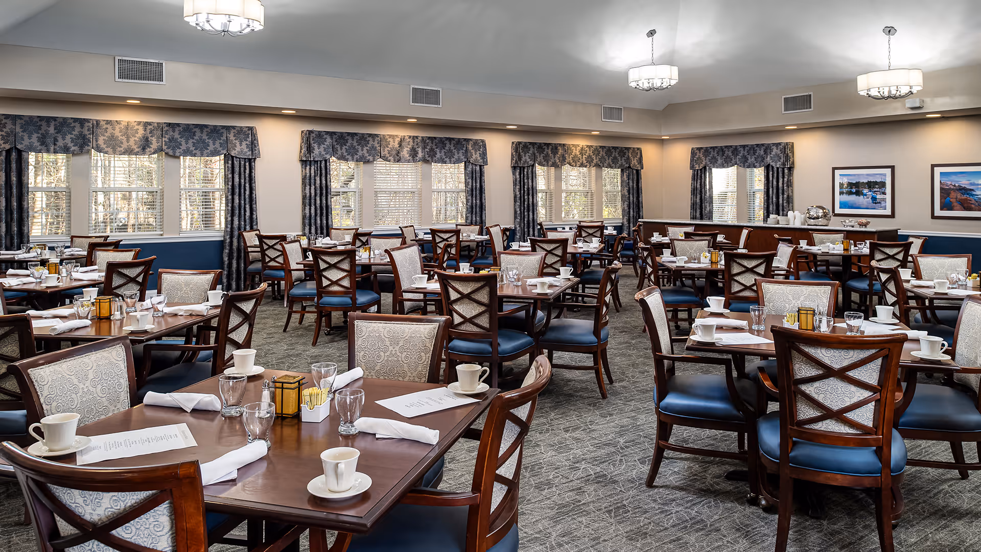 A spacious dining room with multiple wooden tables and chairs arranged neatly. Each table is set with white cups, glasses, napkins, and menus. The room has large windows with blue patterned curtains allowing natural light to enter. The walls are decorated with framed pictures, and there are ceiling lights providing additional illumination.