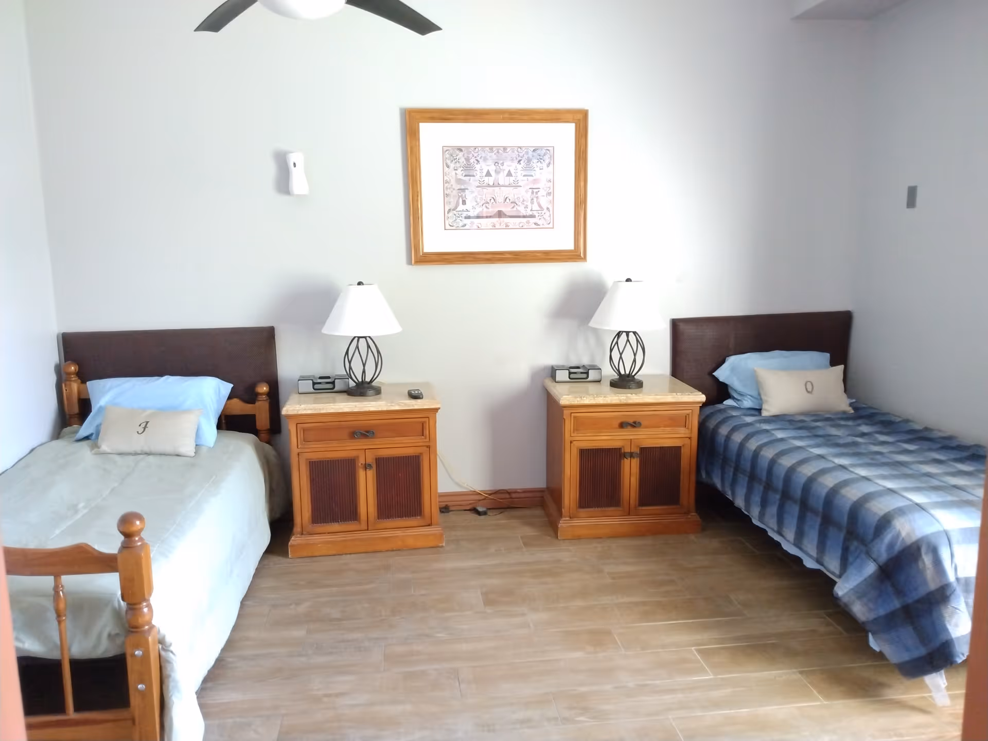 A simple bedroom with two single beds separated by two wooden nightstands with lamps. Each bed has a pillow with an initial, one with 'J' and the other with 'Q'. A framed picture hangs on the wall above the nightstands, and a ceiling fan is visible at the top.