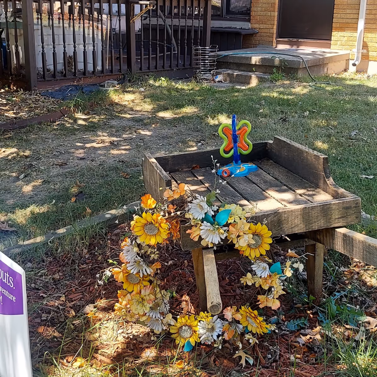 Outdoor garden area with a small wooden wheelbarrow decorated with a colorful floral wreath and a butterfly-shaped toy on top. In the background, there is a porch with a railing, some steps leading to a door, and a garden hose on the ground.