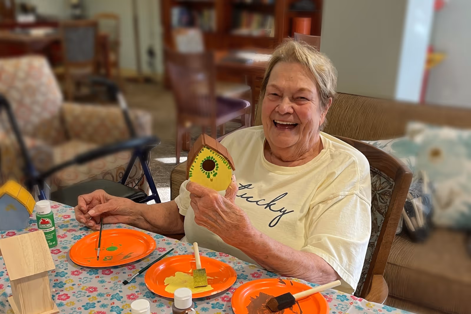 An elderly woman sitting at a table in a living room area, smiling and holding a small painted birdhouse. The table is covered with a floral tablecloth and has paint supplies including brushes, paint bottles, and orange plates with paint on them. In the background, there are chairs, a walker, and shelves with books and other items.