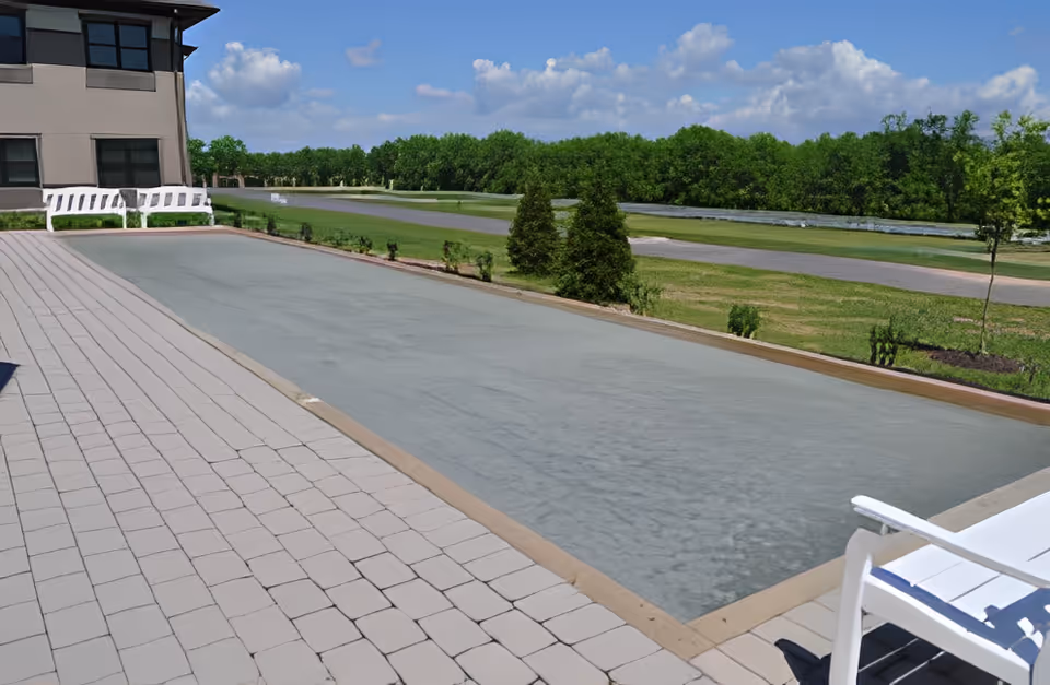 Outdoor patio area with a long rectangular water feature or shallow pool, surrounded by paved stone flooring. There are white benches and chairs placed around the patio. In the background, there is a grassy area with trees and a clear blue sky with some clouds.