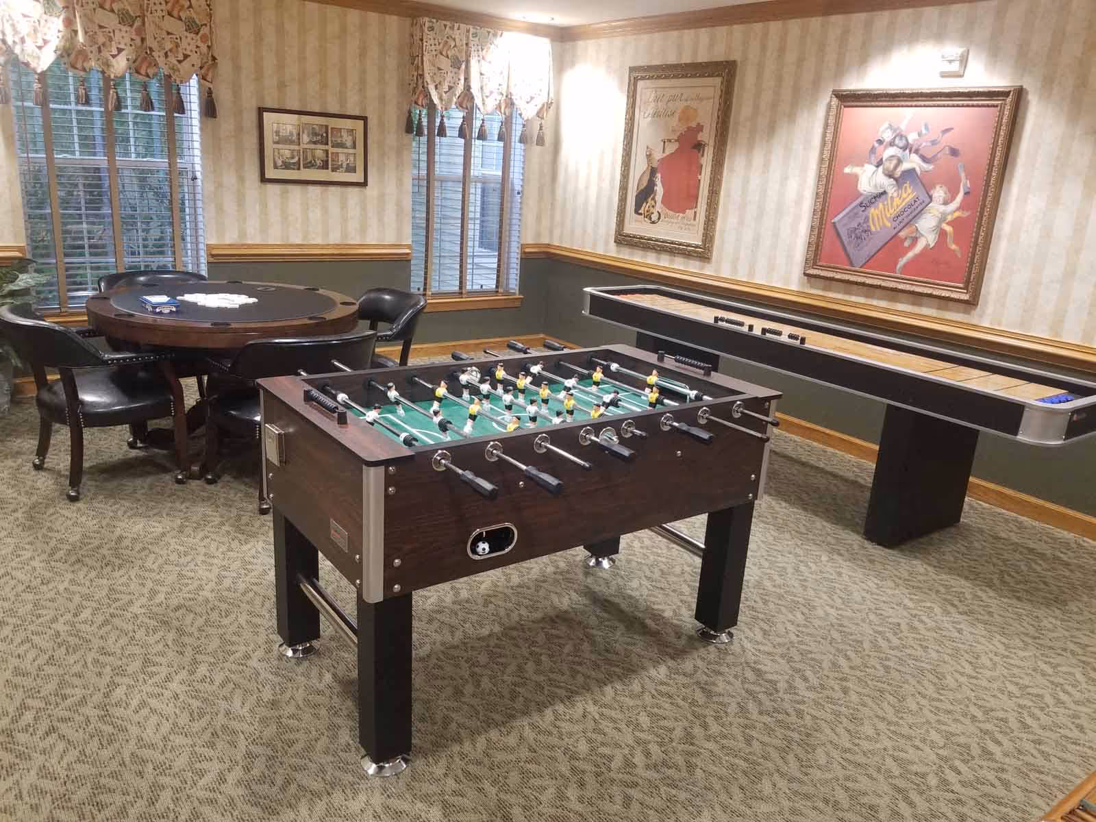 A game room with a foosball table in the foreground, a shuffleboard table against the wall, and a round poker table with chairs near two windows with patterned valances. The walls have striped wallpaper with framed vintage-style posters.