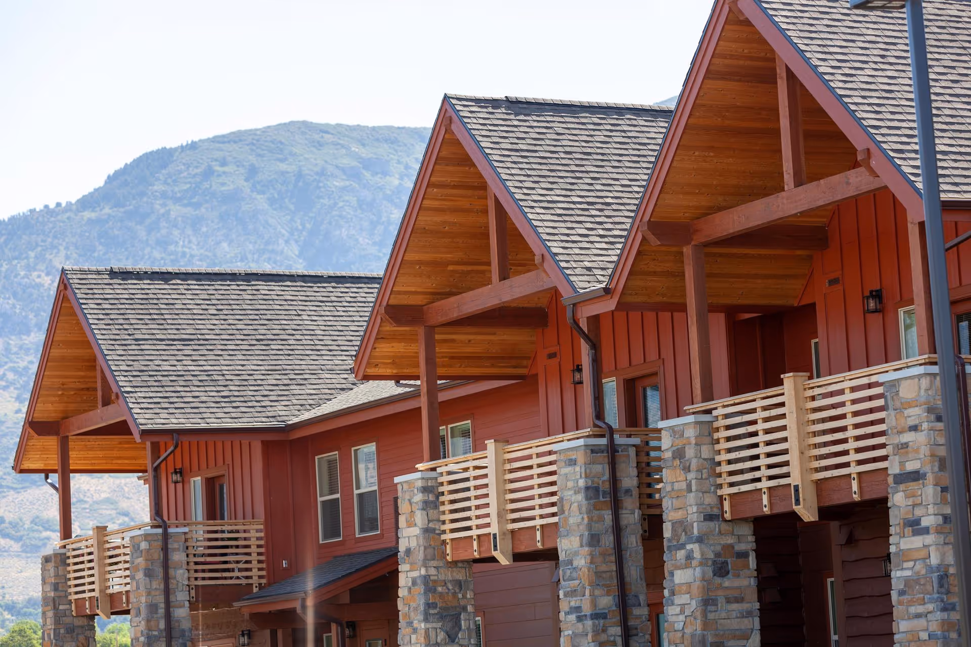 Exterior view of a red building with multiple peaked roofs and wooden balconies supported by stone pillars, set against a backdrop of green mountains under a clear sky.
