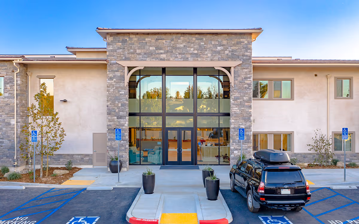 Entrance of a modern two-story senior living building with a stone facade, large glass doors, planters, and a parked SUV in handicapped parking spots.