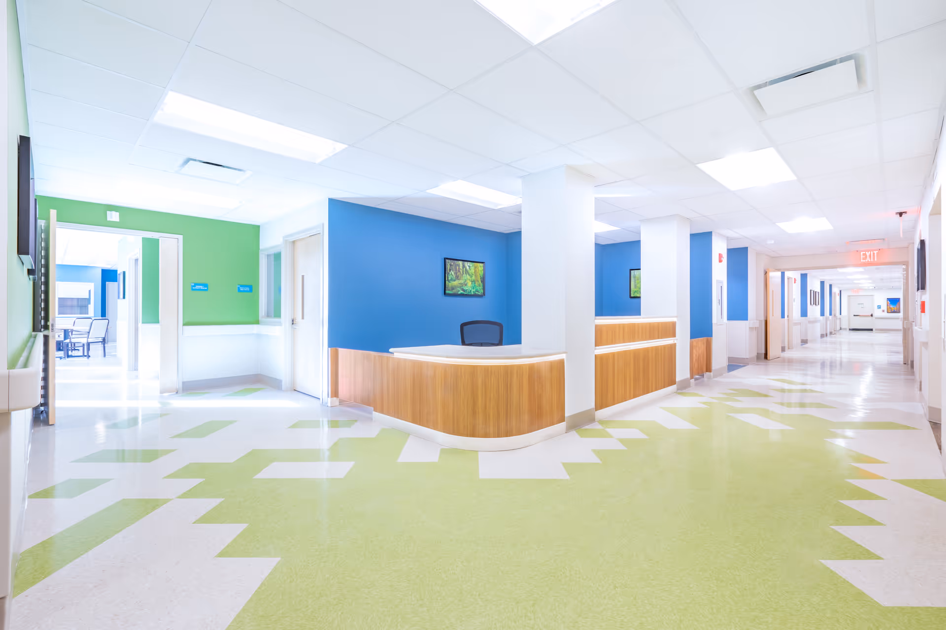 Bright and clean hallway in a senior living facility with a reception desk featuring blue walls and wooden paneling. The floor has a green and white geometric pattern, and there are multiple doors and an exit sign visible down the corridor.