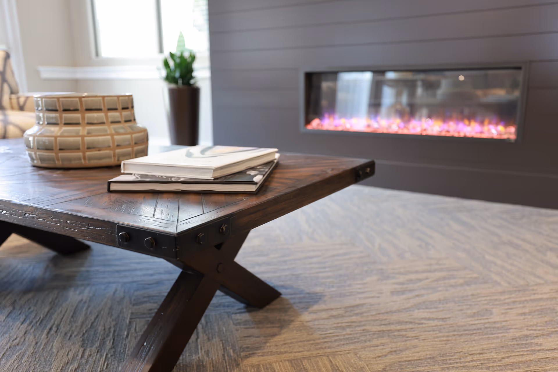 Close-up of a wooden coffee table with two books and a decorative ceramic vase on top, placed on a carpeted floor in front of a modern electric fireplace with purple flames. A potted plant is visible in the background near a window.