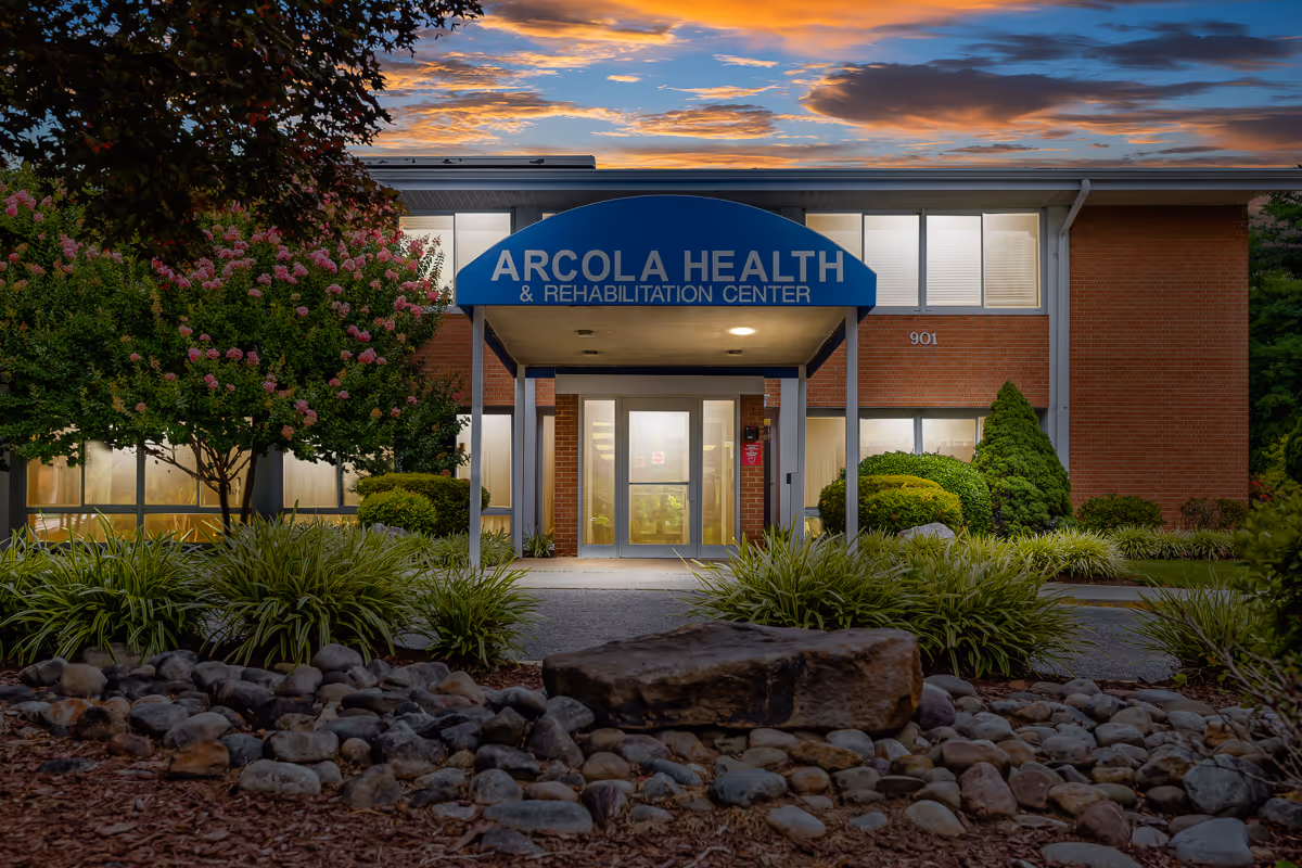Front exterior view of Arcola Health & Rehabilitation Center building at dusk with a blue awning over the entrance, surrounded by landscaped bushes, trees, and rocks.