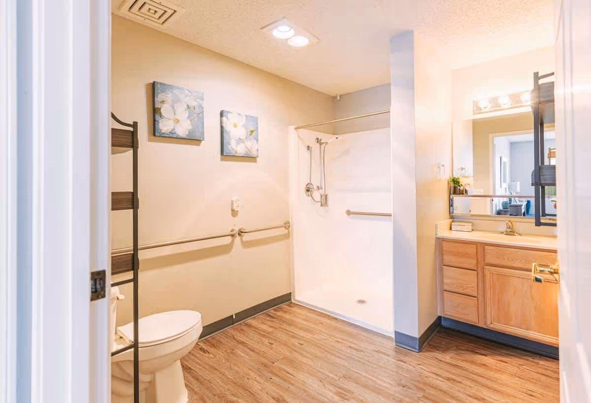 A bright and spacious bathroom featuring a walk-in shower with grab bars, a toilet with a grab bar beside it, a wooden vanity with a sink and mirror, and two floral paintings on the wall. The floor has wood-like vinyl flooring.