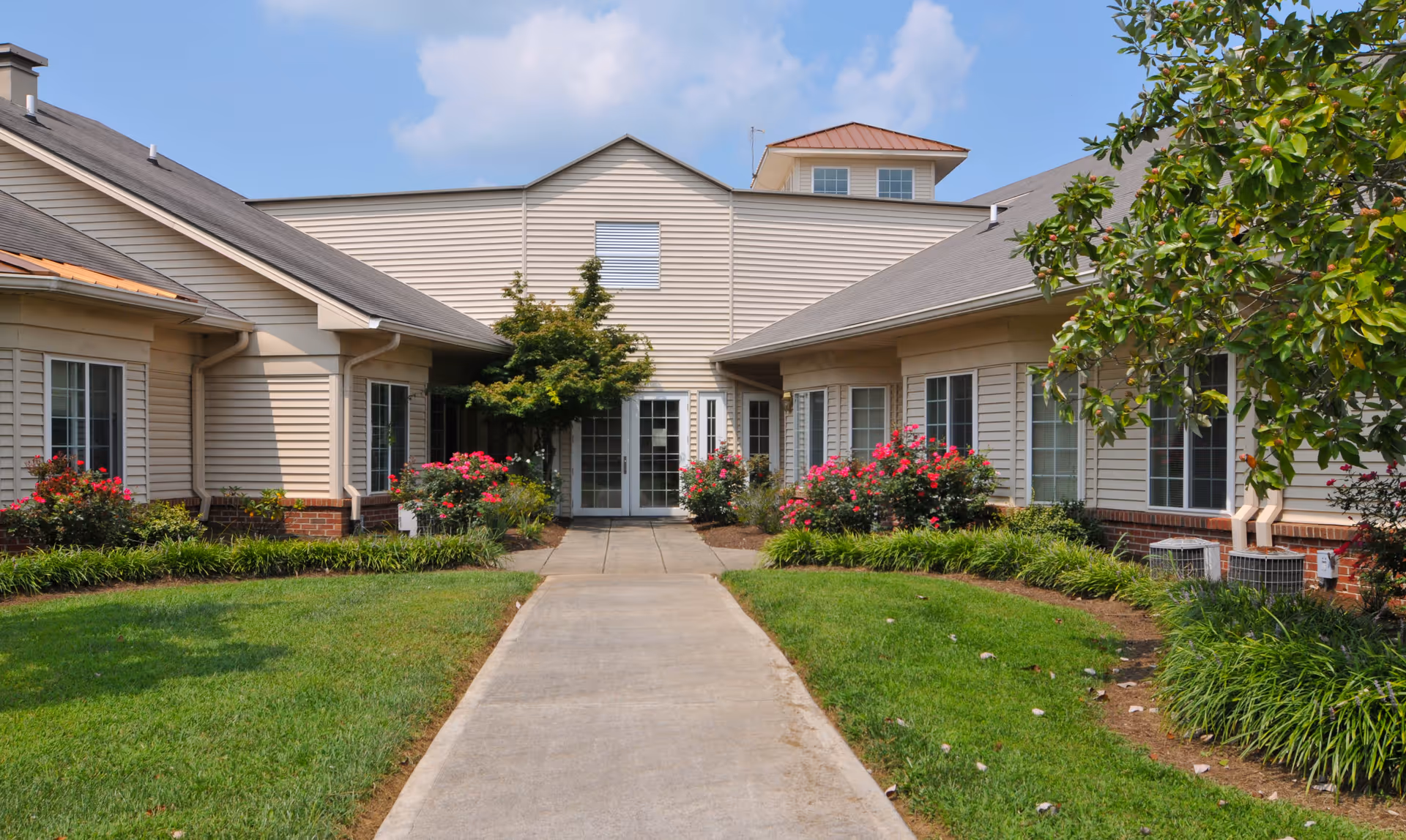 Walkway leading to the front entrance of a beige, single-story building with landscaped lawns and flowering shrubs.