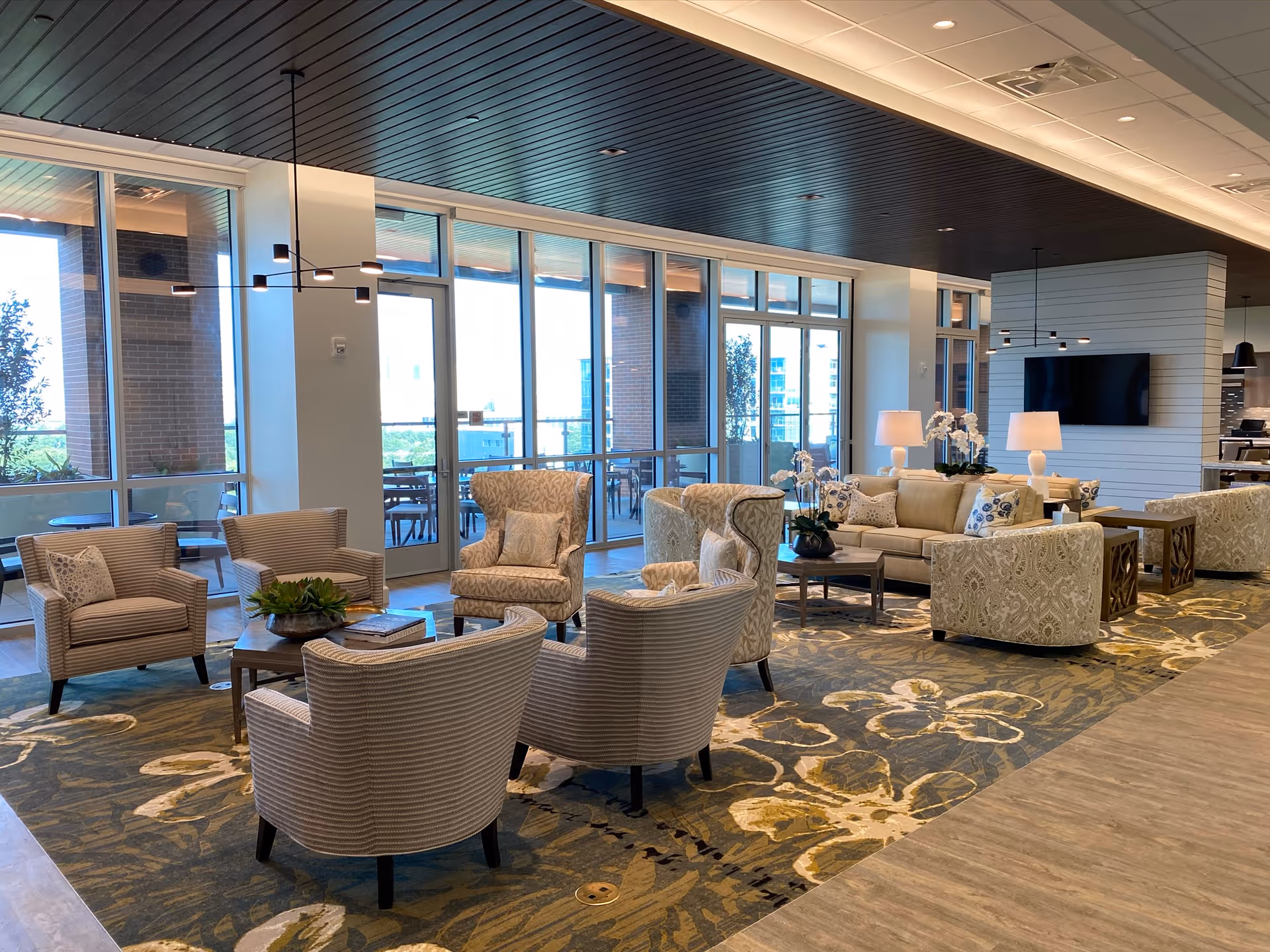 Spacious, well-lit communal living room with sofas, armchairs, coffee tables and large windows overlooking an outdoor patio.