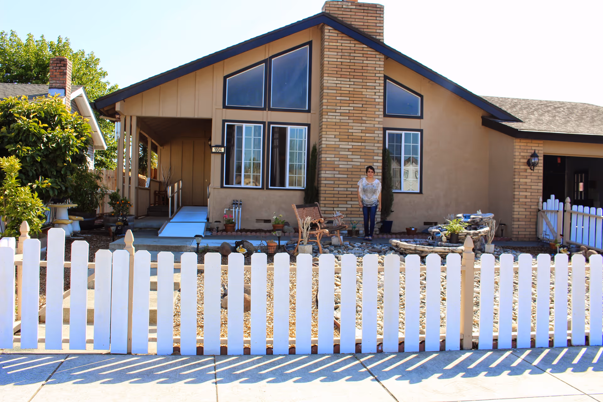 Front of a single-story house with a white picket fence, rock landscaping, and a person standing near the entrance.