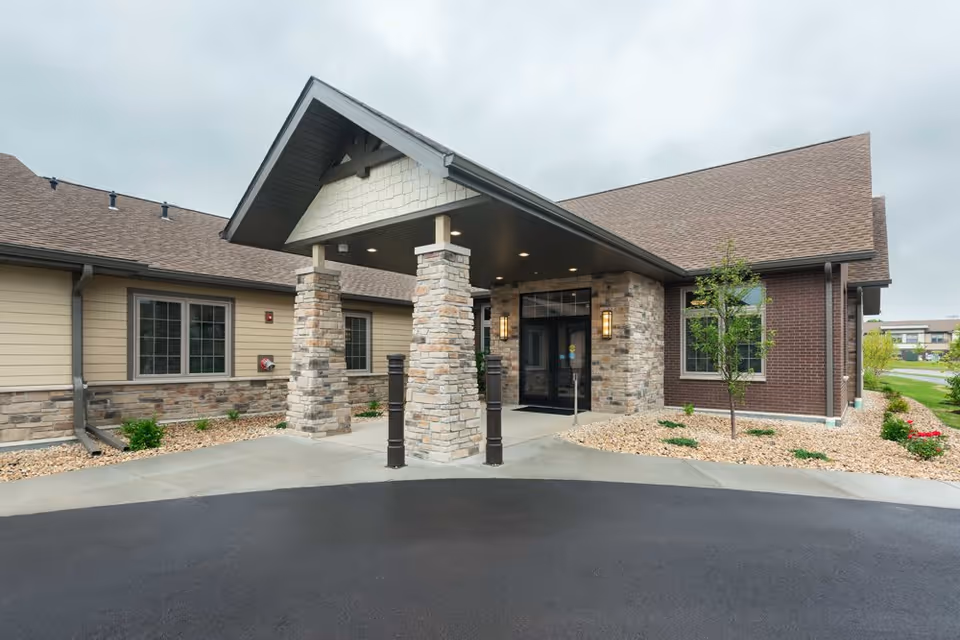 Entrance of The Legacy at Noel Manor facility showing a covered driveway with stone pillars, beige and brown exterior walls, windows, small landscaped areas with rocks and plants, and a paved driveway under an overcast sky.