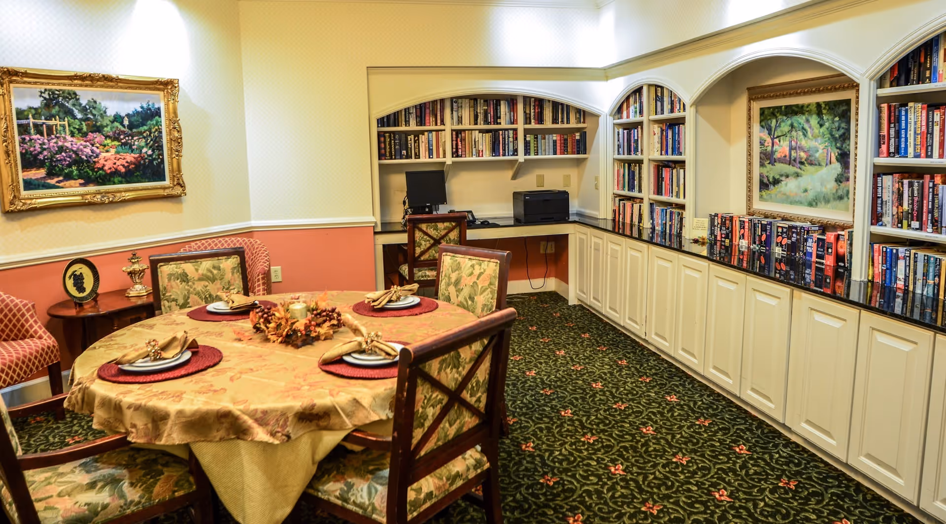 A cozy room with a round table covered with a gold tablecloth and set with four place settings, each with a plate, napkin, and napkin ring. The room has floral patterned chairs around the table, a green carpet with a floral design, built-in bookshelves filled with books, a computer and printer on a desk in the corner, and framed landscape paintings on the walls.