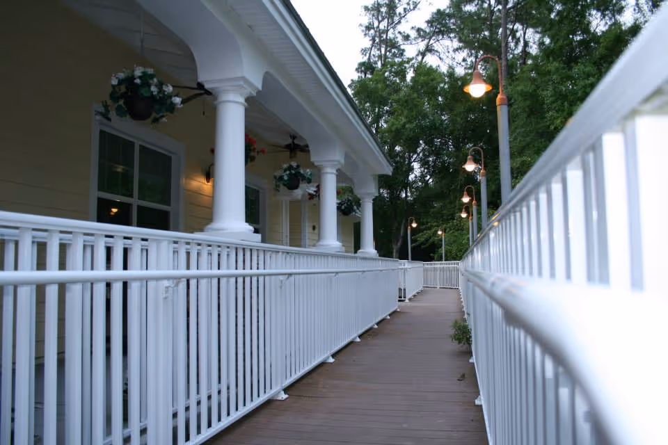 A wooden ramp with white railings leading to a building porch with white columns, hanging plants, and lampposts amid trees.
