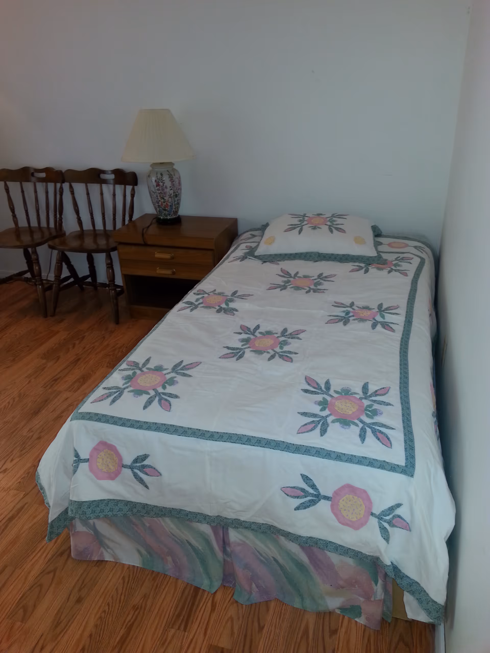 A small bedroom with a single bed covered in a white quilt with floral patterns in pink, yellow, and green. Next to the bed is a wooden nightstand with two drawers and a decorative lamp with a floral design. Two wooden chairs are placed against the wall beside the nightstand. The floor is wooden, and the walls are plain white.