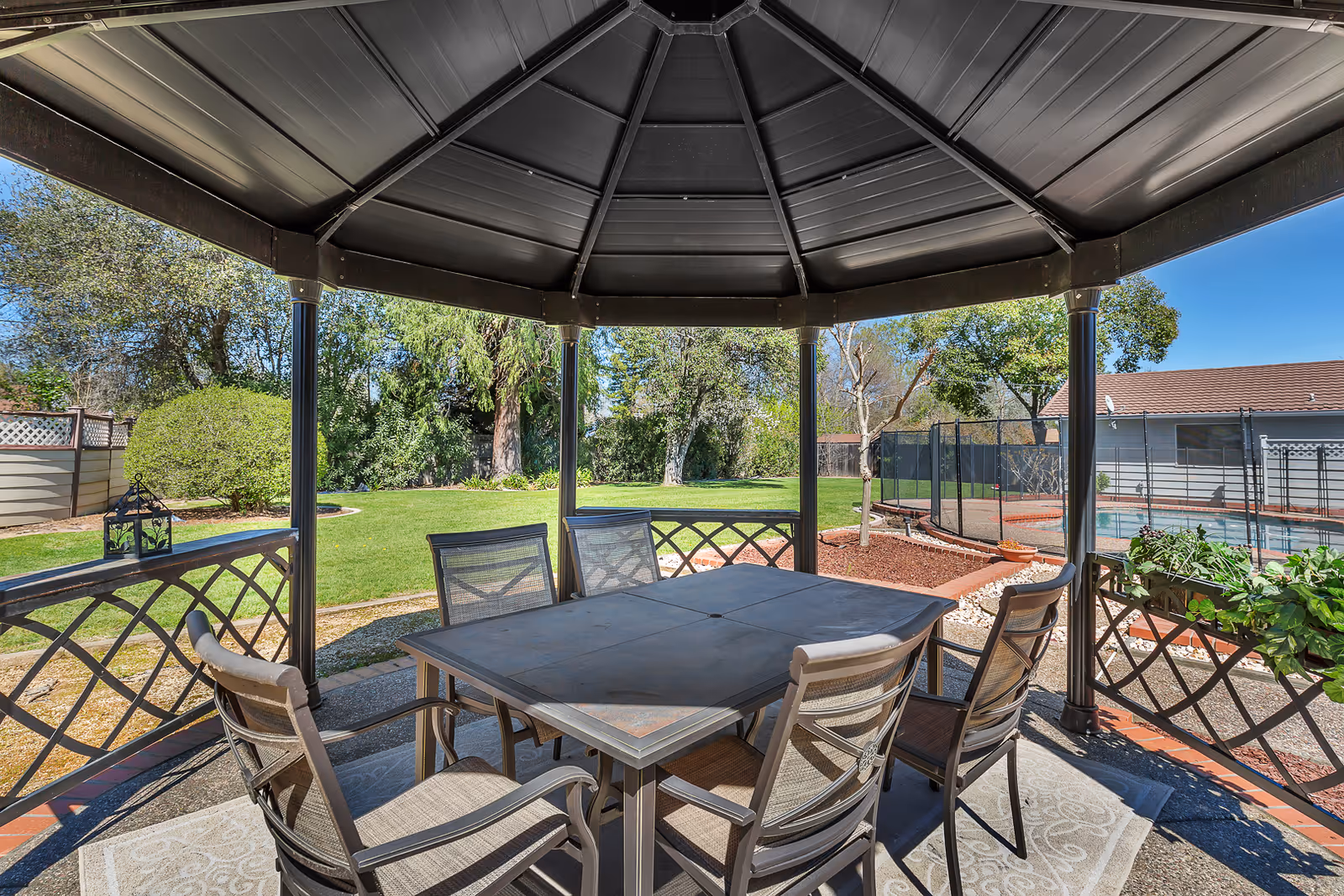 Outdoor gazebo with a metal roof covering a square table and six chairs on a patterned outdoor rug. Surrounding the gazebo is a well-maintained lawn, trees, and a fenced swimming pool area with a building in the background under a clear blue sky.