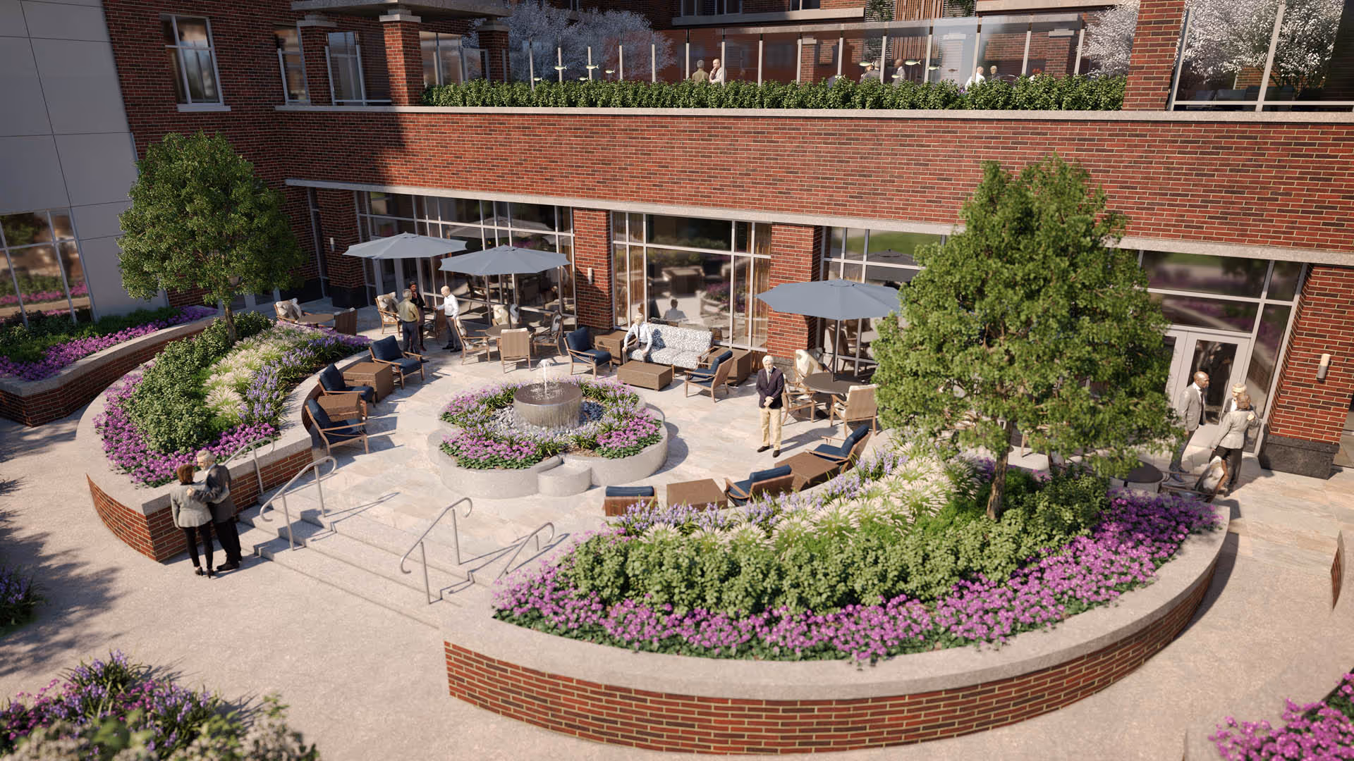 Outdoor patio area at The Providence Fairfax featuring a circular seating arrangement around a central water fountain. The space is surrounded by curved brick planters filled with green shrubs and purple flowers. Several people are standing and sitting under large umbrellas near the building's glass doors and windows. Trees and additional landscaping enhance the garden-like atmosphere.