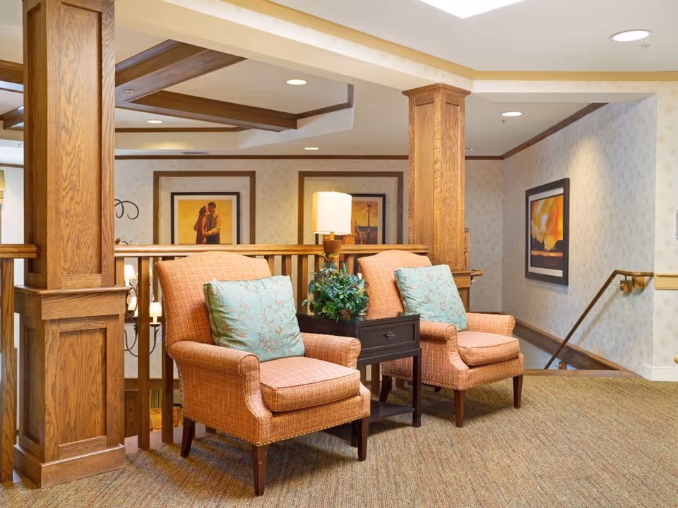 Two cushioned armchairs with patterned upholstery and light blue decorative pillows are placed side by side in a cozy indoor seating area. Between the chairs is a small dark wooden table with a lamp and a green potted plant. The space features wooden railings and columns, beige patterned wallpaper, framed artwork on the walls, and carpeted flooring. A staircase with a wooden handrail is visible to the right.