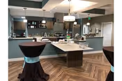 Modern communal kitchen and dining area with a central island, stainless appliances, pendant lights, and two draped high-top tables.