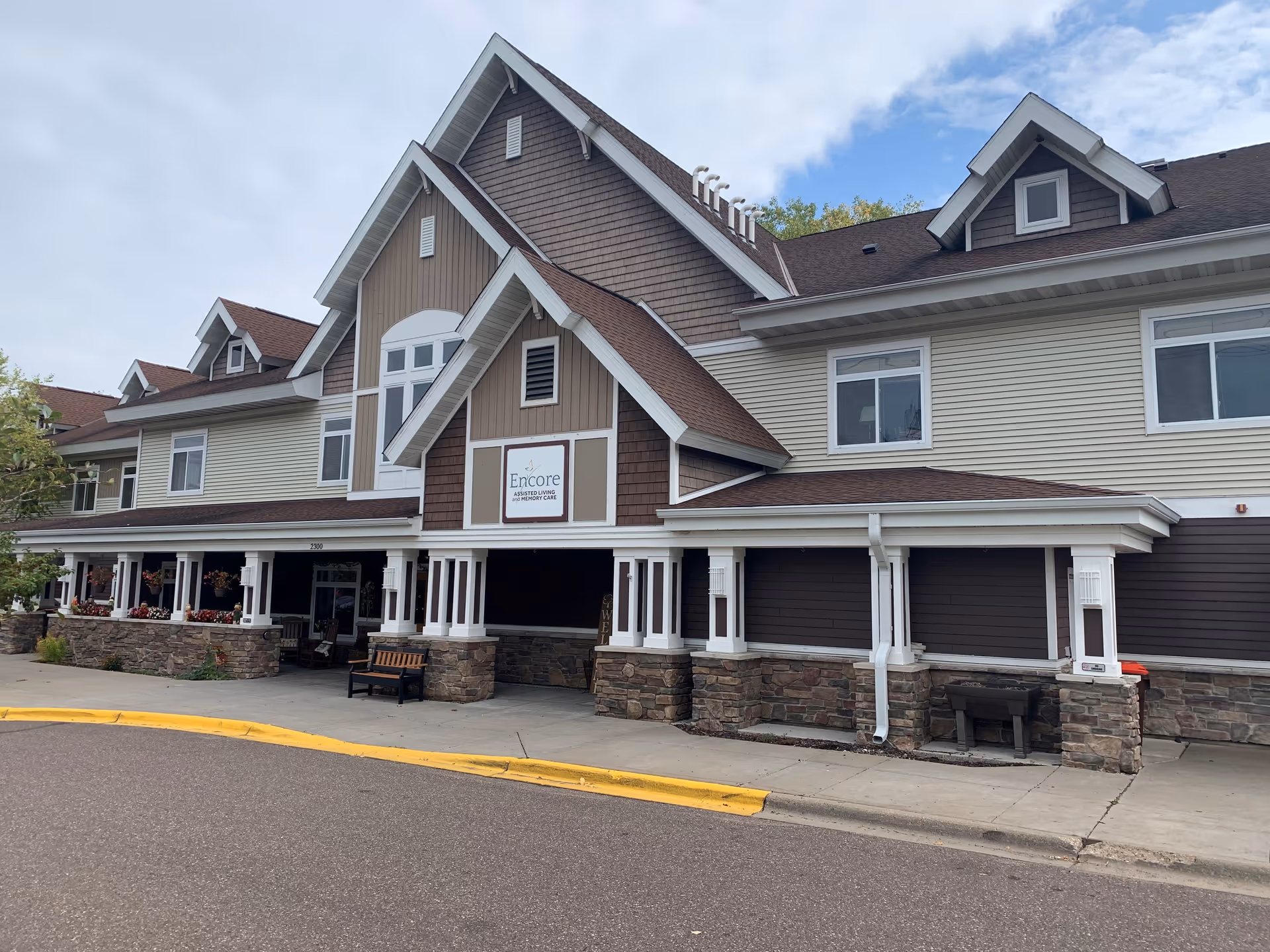 Exterior front view of Encore Assisted Living and Memory Care at Maplewood, showing a large building with brown and beige siding, multiple windows, a covered entrance with white columns, stone accents, and a bench near the entrance.