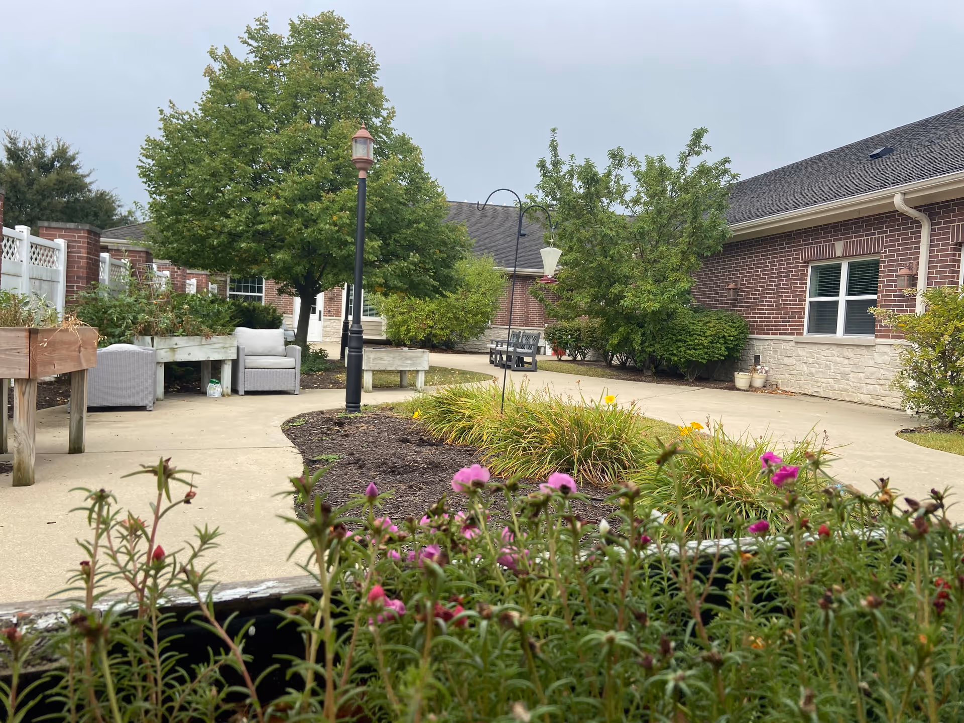 Outdoor garden area at Aspen Woods of Vernon Hills featuring a paved walkway, flower beds with blooming pink and yellow flowers, green trees, shrubs, and outdoor seating including cushioned chairs and benches. The building with brick and stone exterior walls is visible in the background under a cloudy sky.