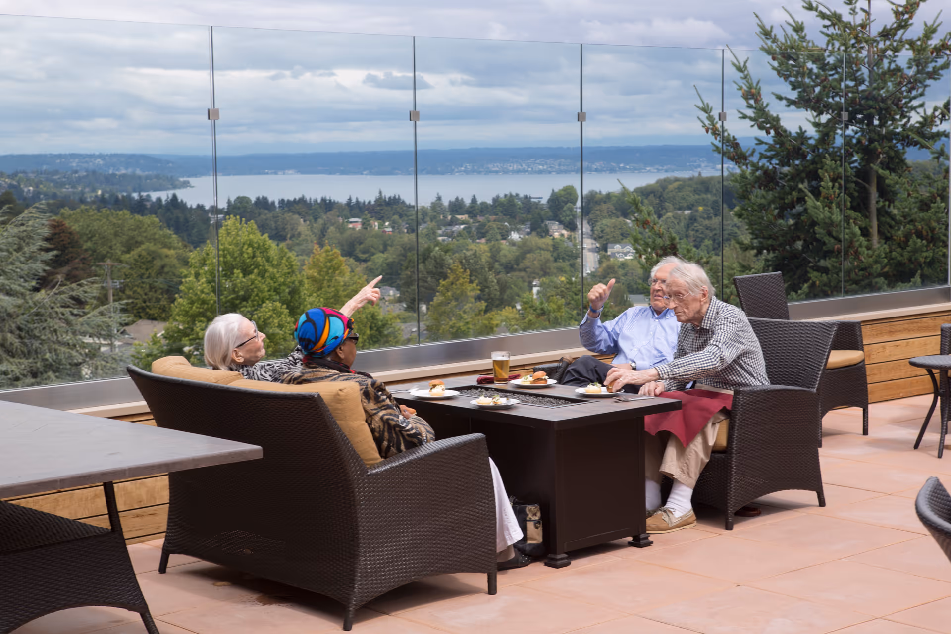 Four elderly people sitting outdoors on a patio with wicker furniture, enjoying food and drinks around a fire pit table. They are overlooking a scenic view of trees, houses, and a body of water in the distance under a cloudy sky.