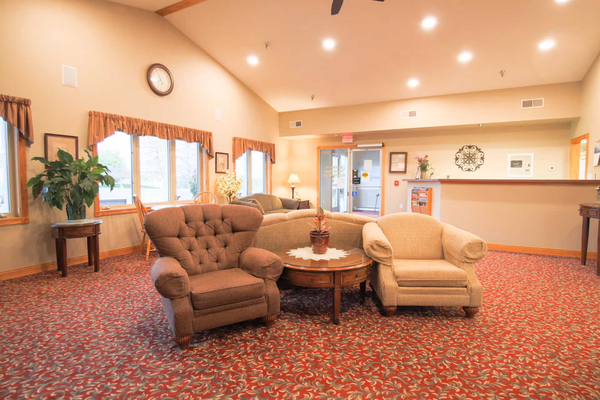 Spacious assisted living common lounge with armchairs around a coffee table and a reception desk in the background.