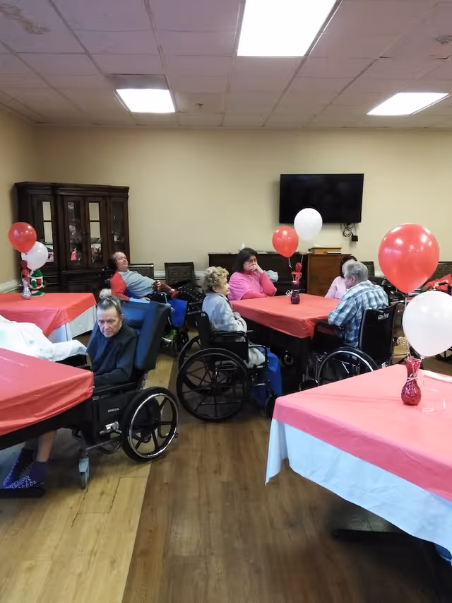 A group of elderly people, some in wheelchairs, sitting around tables covered with red and white tablecloths in a common room decorated with red and white balloons. There is a wooden cabinet and a wall-mounted TV in the background.