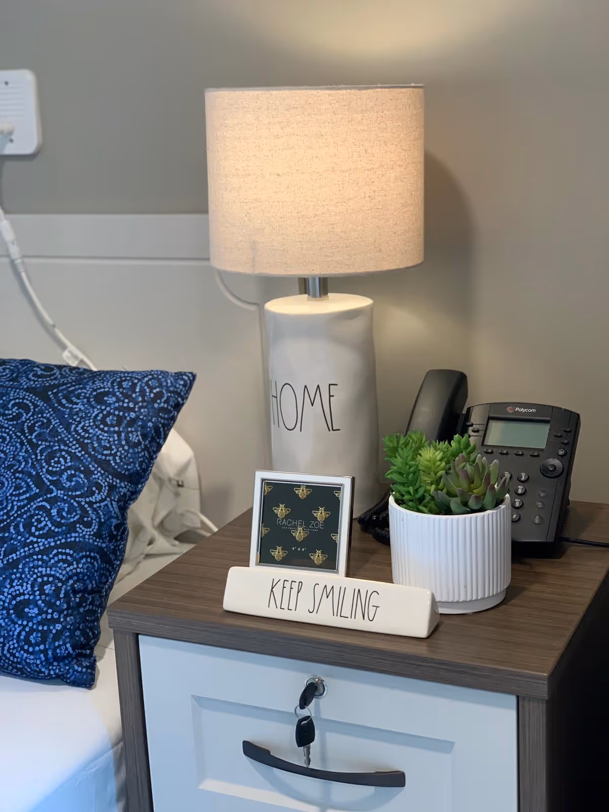 A bedside table with a white drawer and dark wood top, holding a white lamp with the word HOME on it, a small framed picture, a white planter with green succulents, a black office phone, and a decorative sign that says KEEP SMILING. A blue patterned pillow and part of a bed are visible next to the table.