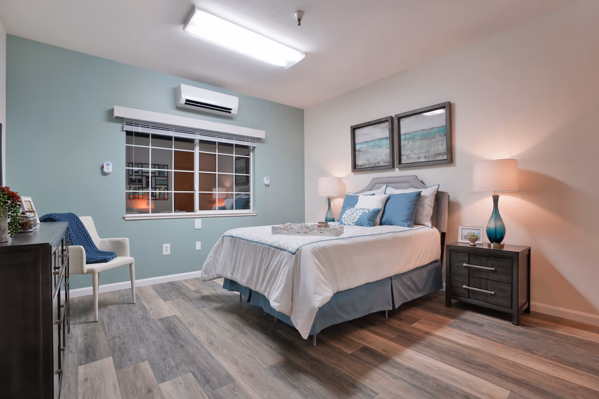 A neatly arranged bedroom with a bed dressed in white and blue bedding, two bedside tables each with a lamp, a chair with a blue throw, a dresser with decorative items, and two framed artworks above the bed. The room has wood flooring, a window with blinds, and a wall-mounted air conditioning unit.