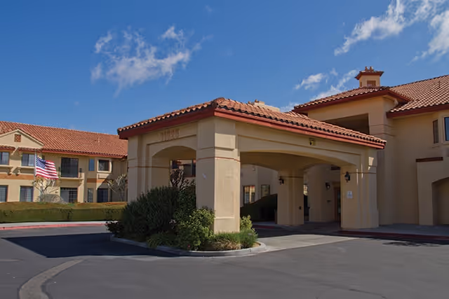 Covered entrance/porte-cochère of a beige stucco senior living building with a red tile roof and an American flag by the driveway.
