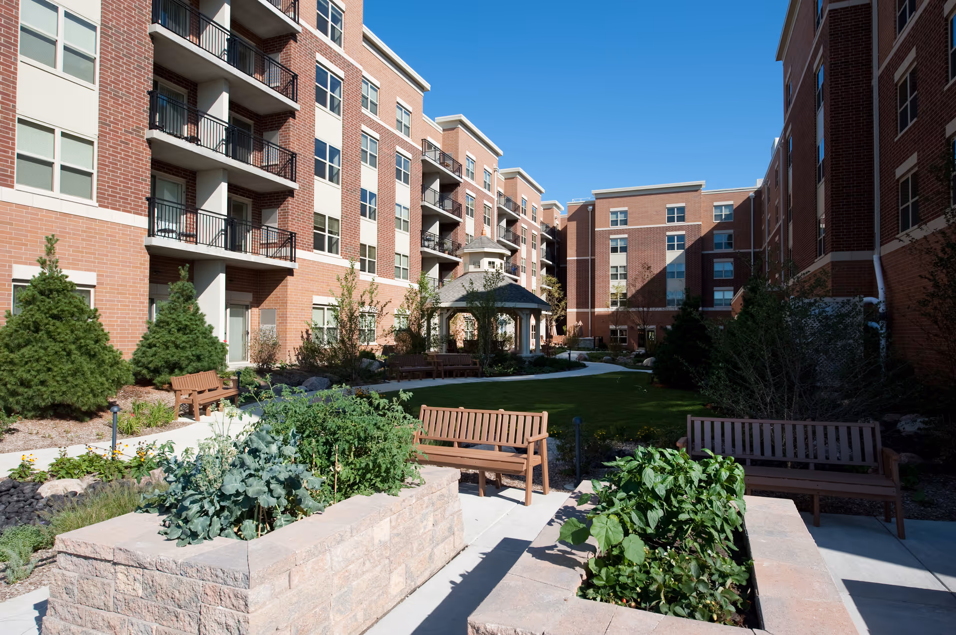 Outdoor courtyard area of a senior living facility with raised garden beds, wooden benches, a gazebo, and multi-story brick buildings surrounding the space under a clear blue sky.