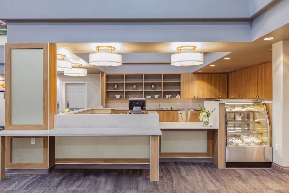 A clean and modern reception or service counter area with light wood cabinetry, white marble countertops, and a glass display case containing pastries or baked goods. The space is well-lit with round ceiling lights and has open shelving with cups and small decorative items in the background.