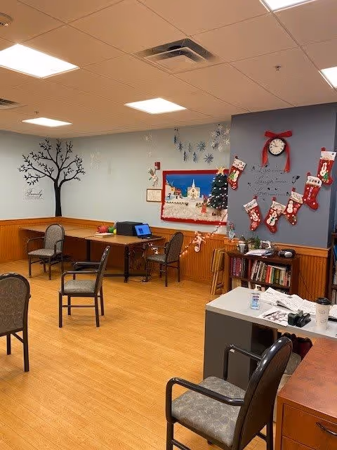 A cozy room with wooden flooring and light blue walls decorated with a black tree wall decal and holiday decorations including Christmas stockings, a small decorated Christmas tree, and a festive wall hanging. The room contains several chairs, a desk with a laptop, a bookshelf with books, and a table with papers and a coffee cup.