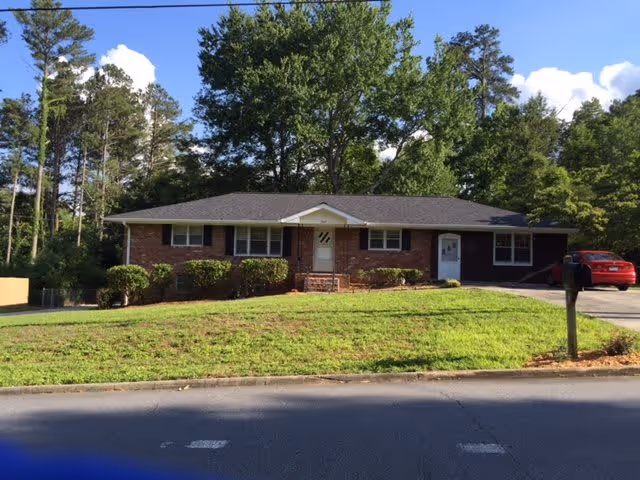 Front exterior of a single-story brick ranch house with a grassy lawn, driveway, mailbox and surrounding trees.